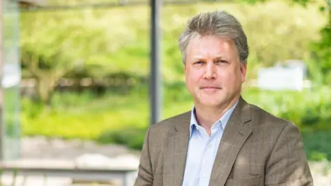 Jeff Dowling/EMBL-EBI The male professor, who has greyish-brown hair, stares at the camera wearing a blue shirt and brown checked suit - there is a blurred green background of trees and bushes behind hime