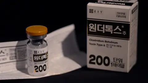 A close-up of a small glass vial and its packaging labelled in Korean script. The box shows the words ‘Clostridium Botulinum Toxin Type A’ with the number 200. The vial has a yellow cap and sits on a folded Korean instruction sheet.