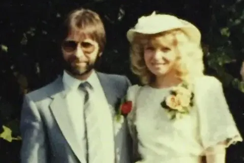 Kate Gilmore Dave, sporting a beard and 80s aviators, in a suit with a blonde-haired Kate next to him in her bridal dress 