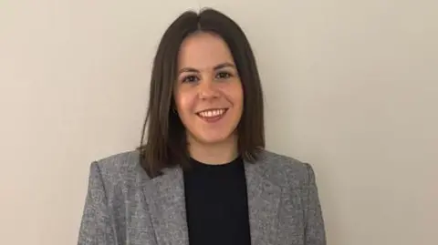 Silvia Prades A woman with brown hair wearing a grey jacket  and a black top smiles in front of a plain wall.