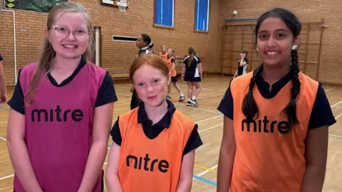 Three Year Seven girls are in a school sport hall.  They are smiling at the camera. They are wearing navy short-sleeved uniform T-shirts and coloured netball bibs. A game of netball is taking place in the background. 