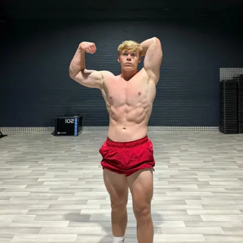 George Holland George Holland a teenager poses for the camera in a body-building stance, wearing red shorts and flexing.