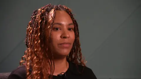 BBC Lauren Caulfield, who has brown, braided hear. She wears a neutral expression as she talks to a journalist.