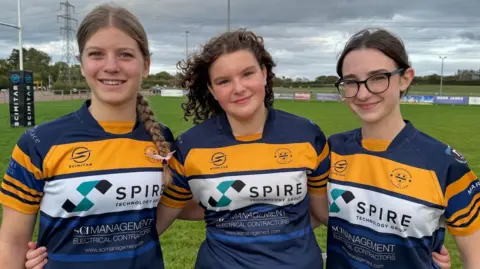 BBC The three teenagers are standing on a rugby pitch with their arms around each other.  They are smiling and wearing blue and gold Worcester rugby club shirts. 