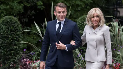 Getty Images French President Emmanuel Macron and First Lady Brigitte Macron arriving in Downing Street in July. The image depicts the couple walking arm in arm. Mr Macron is dressed in a dark blue suit, white shirt, and dark tie. Mrs Macron is wearing a light-colored, double-breasted jacket suit. They are surrounded by greenery, including bushes and plants with long leaves