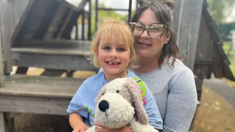 Holly wearing a grey top and glasses with her daughter Ayda sitting on her lap holding a cuddly toy dog 