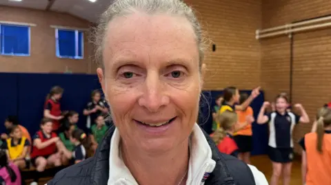 Liz Brown is wearing a white sports top and navy Gillet.  She's smiling at the camera. She's in a school sports hall and there are students laughing behind her.