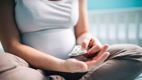 Getty Images A pregnant woman sits cross-legged on a bed and holds a blister pack of medication. There is an out-of-focus white cot in the background. 