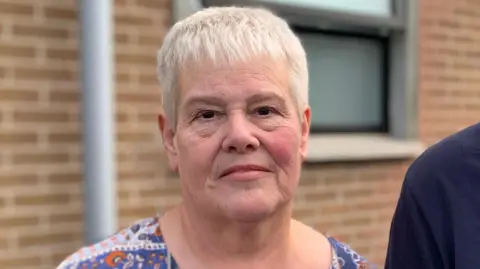BBC A woman with short white hair and brown eyes looks into the camera. Cut off at the shoulder you can see she wears a blue, orange and white coloured top. A mans shoulder can be seen to her left, he wears a navy top. Behind her, out-of-focus is a red bricked wall. A partially opened translucent window is positioned over her left shoulder. 