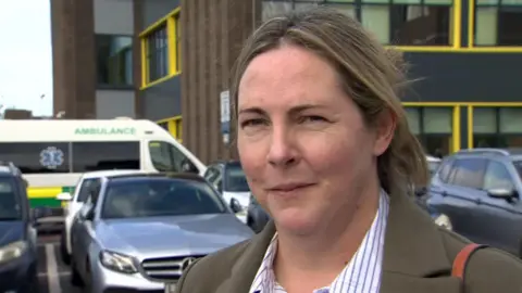 A woman stares at the camera. She has long blonde hair, and is wearing a blue and white striped shirt and a green blazer. Behind her is a buildings with cars and an ambulance parked in front of it.