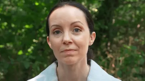 BBC Morning Live Stef Reid standing outside presenting for BBC Morning on a sunny day. She has brown hair and is wearing a green jumper