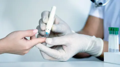 Getty Images Close up of hands as patient undergoes finger prick test administered by a member of hospital staff who is wearing white plastic gloves.  