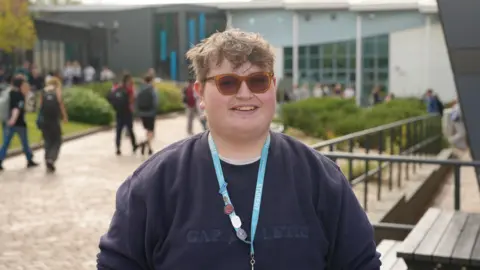 17-year-old student Sam stands and smiles in the courtyard of his FE college. He is wearing sunglasses and his college lanyard