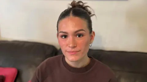 Molly looks at the camera. She has dark hair pulled back behind her head. She is wearing a reddy-brown jumper.