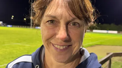 Nicky Crawford is standing next to a floodlit rugby pitch.  She's wearing a hooded training top and smiling. 