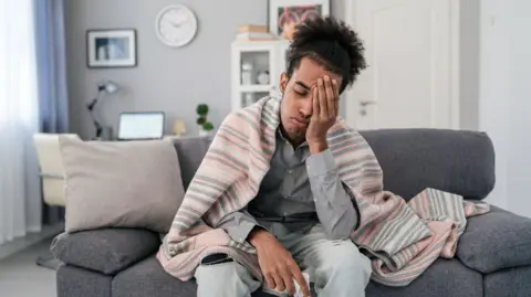 Getty Images A young man, who looks unwell, sits on his sofa, wrapped in a blanket, resting his head in one hand, while holding a tissue in the other. 