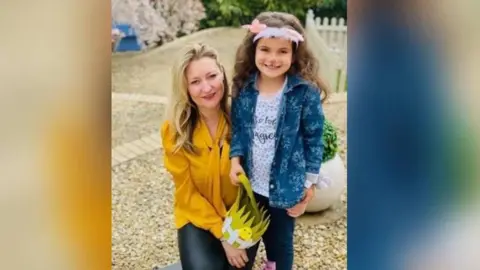 Zoe Hoes A woman who has blonde hair wearing a yellow top. She is holding a young girl around her waist as the pair smile for a photo. The girl has cat face paint on her face and is wearing a blue denim jacket and white top. They are stood in a stone area.