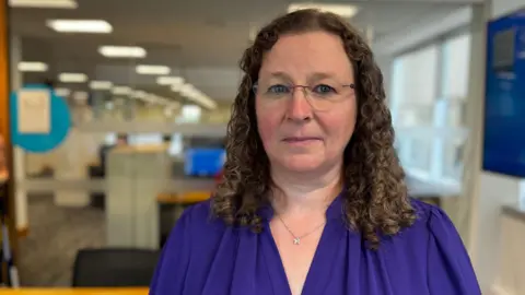 A woman with dark brown shoulder length curly hair and rimless glasses stands  with an open plan office in the background. She is wearing a dark purple shirt.