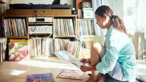Getty Images A woman kneels on the floor surrounded by music albums in a room with shelves filled with record albums.