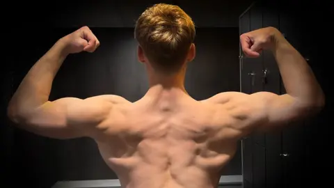George Holland A teenage boy flexing his muscles and showing his back.