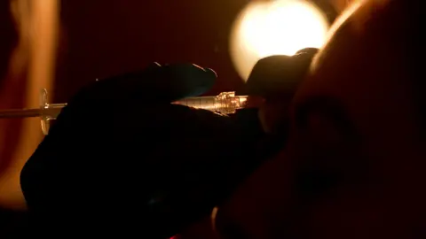 A silhouetted hand prepares a syringe close to a woman’s face under a bright light, representing the risks of unregulated cosmetic injections