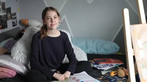 Avalyn, a 16 year old with long Covid, sits on her bed smiling propped up with cushions, with the books she used for GCSE revision open alongside her. 