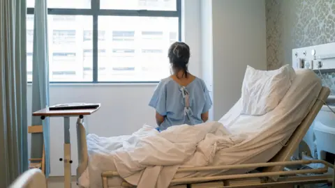 Getty Images An anonymous woman sitting in an upright hospital bed. She is looking out of a window and her back is facing the camera. She is wearing a blue hospital gown.