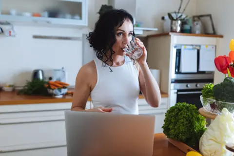 Getty Images A woman in a white top drinks water in a kitchen surrounded by fresh vegetables and a laptop