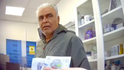A man with grey hair stands in a clinic office, looking towards the camera. Shelves with medicine boxes and bottles are behind him. In the foreground, a hand is holding out cash.