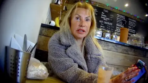 A woman with blonde hair, wearing a grey coat, sits at a café table holding a phone. A drink is on the table in front of her, and shelves with jars and lights are visible in the background.
