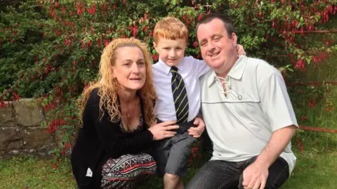 McGerr Family Smiling parents and their young boy smile for the camera on the boy's first day of school - the mum has strawberry blonde hair and is wearing a black top and multi-coloured dress. The dad is wearing a light top, jeans and has close cropped dark hair. The boy has reddish hair, a green tie and grey shorts.