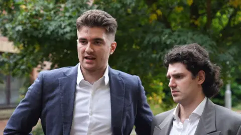 PA Media Two young men stood outside in front of some trees. On the left, a man wearing a blue suit jacket and white shirt. he has short brown hair. On the right, a man in a grey suit jacket and white shirt with long curly black hair
