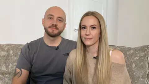 BBC Tom and Becky Williams are sitting on a large, neutral coloured sofa. Tom has a tattoo on his arm and is wearing a dark grey top, he is bold and has a beard. Becky is wearing a neutral coloured top, she has blonde hair and together they are looking at the camera. Being them is the door to their living room.