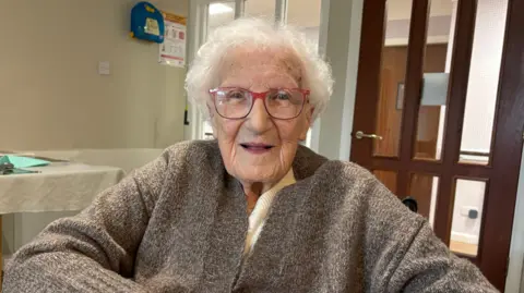 BBC An elderly woman, 100-year-old May O'Shea, smiles into the camera. She has  short, white curly hair, blue eyes, red glasses and is wearing a brown cardigan.