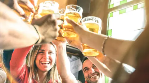 Getty Images A young woman and a man smile as a large number of arms holding pint glasses of lager or beer are held above their heads, as if they are saying cheers