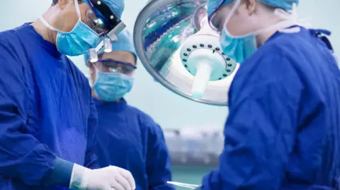 Getty Images Surgeon with a head light on and blue mask performs a surgery out of sight of the camera. There are two other surgeons dress in blue robes beside him. 