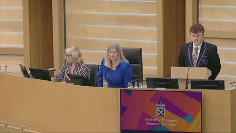 Scottish Parliament A teenage boy standing up behind a podium in a parliamentary building, wearing a black school uniform with purple trim and tartan tie. Two women are sitting along from him, one wearing a blue suit and the other in a multi-coloured top.  The walls and decor of the room are beige.