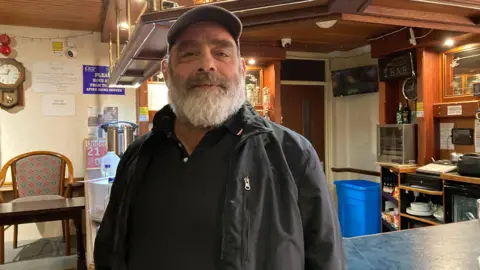 BBC A man with a beard and cap, and wearing a black shirt and black jacket. Stands in front of a bar and smiles slightly.