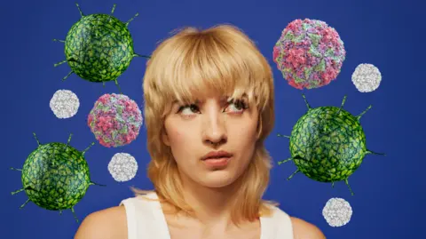 Getty Images A person with blonde hair, dressed in a white top, stands against a blue backdrop. Surrounding them are floating, orb-like structures resembling virus particles.