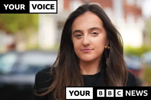 BBC A woman with flowing long brown hair, wearing a black jacket and black top looks pensively into the camera. Behind her are several cars out of focus. Overlaid on top of the photo is a black and white graphic reading 'your voice, your BBC News' in caps.