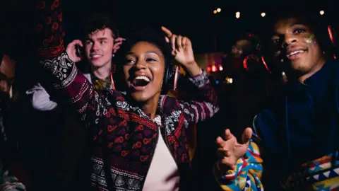Getty Images Young people dance and raise their arms in the air while wearing headphones, in a dark place with a trace of some lights in the background