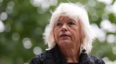 PA Media Women with white hair looking to the side of the camera, upwards. She is wearing a black coat. In the background the leaves of a tree, out of focus.