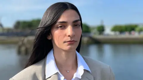 BBC Head and shoulders shot of Ellie Wilson with a river in the background, which is out of focus. She wears a white shirt and an oversized grey blazer. She has long straight black hair.