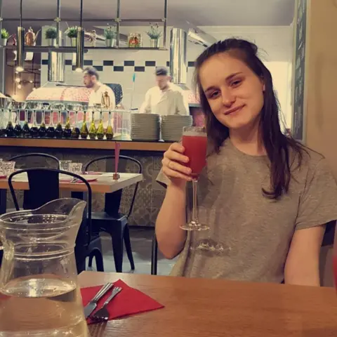 Family handout Cerys Lupton-Jones sitting in a restaurant. She is smiling at the camera and holding up a tall wine glass with a red drink in it. She is wearing a grey short and has long brown hair over her shoulders. Behind her two chefs can be seen working in an open kitchen