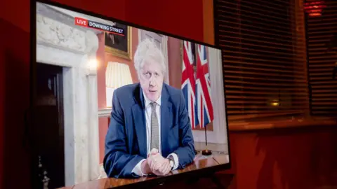 Getty Images A photo of a TV screen in a living room shows Boris Johnson giving a briefing live from Downing Street