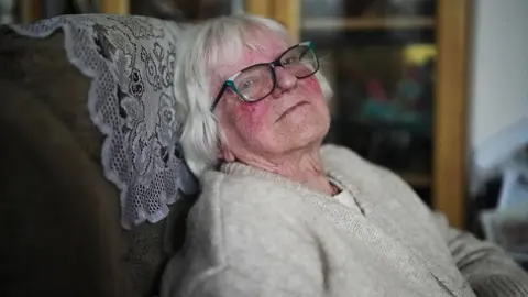 Jean, an elderly person with white hair is seated in a cushioned chair with a patterned fabric cover. The person is wearing a light beige knitted sweater. In the background, there is a wooden cabinet with glass doors displaying various items.