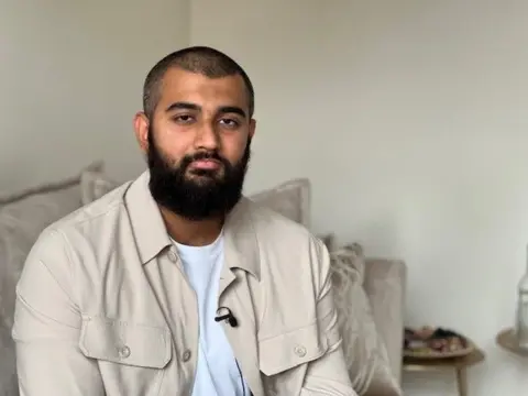 A man with shaved dark hair wears a beige over-jacket with a white t-shirt underneath
