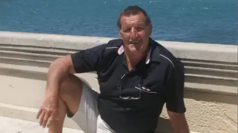 Family photo Man wearing a dark polo shirt and white shorts sitting on a concrete bench/wall next to the sea. It is sunny and he is tanned. 