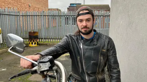 BBC News A young man with long brown hair and a brown beard, wears a black leather jacket and a black back-to-front cap, as he sits on a motorbike in a yard.