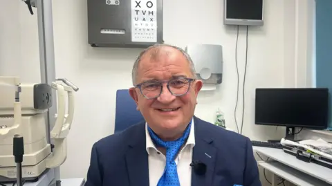 A man wearing a blue suit and blue neck tie is smiling at the camera. Behind him is some medical equipment. 